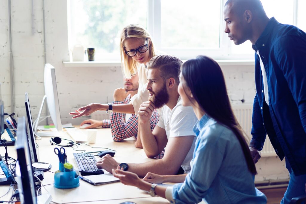 Group of Young Coworkers Sitting Together at Table and Working on Laptops in Modern Office.