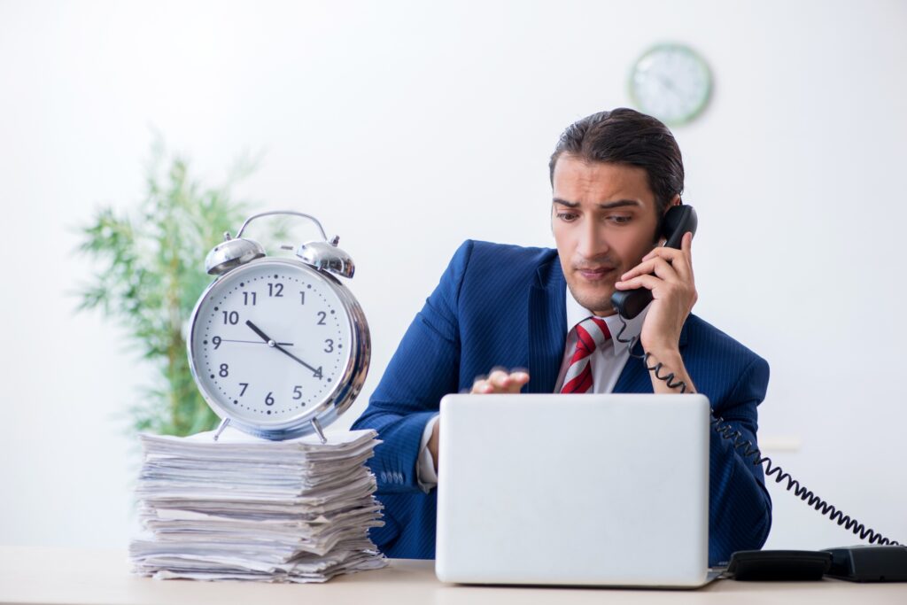 Young male businessman sitting in the office with a stack of papers and a clock on top of it.